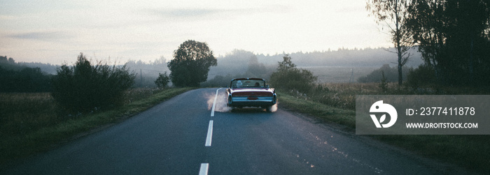 A retro car drives along the road at dawn. The convertible drives off into the distance on a deserted road