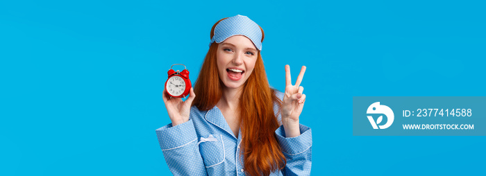 Sassy and cheeky confident redhead caucasian female in nightwear, sleep mask, showing red alarm clock and peace sign, smiling carefree, always be in time, waking up early, standing blue background