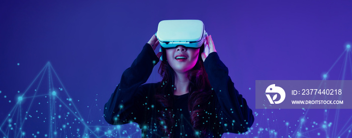 Metaverse technology concept. Excited young Asian woman wearing VR headset happy smile with new experience on futuristic purple cyberpunk neon light banner background.