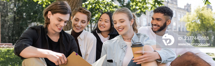 Positive multiethnic students looking at friend with copy book in park, banner.