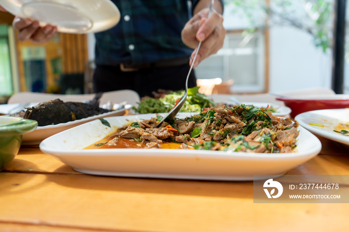 Stir fried duck with basil in square white dish on wood table, is scooped by Asian man with his big spoon for lunch.