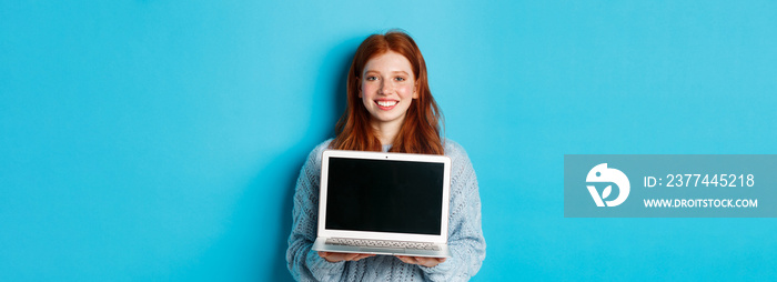 Young smiling woman with red hair and freckles showing computer screen, holding laptop and demonstrate online promo, standing over blue background