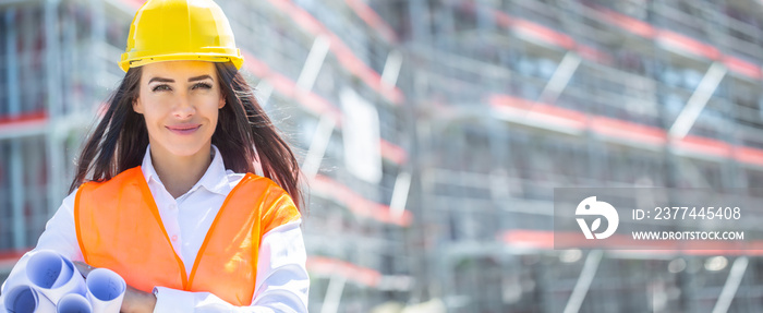 Female architect wearing orange safety vest and a yellow helmet holds rolled construction plans under her armpit in front of the unfinished building