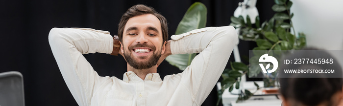 bearded businessman sitting with hands behind head and smiling in office, banner