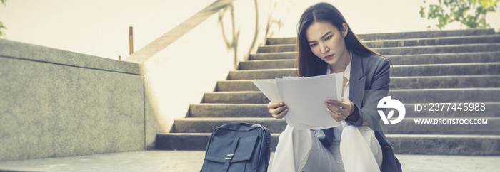 Young woman in a brown suit sits on the stairs holding a white paper, Unemployed business woman sits sadly on the stairs holding the application document, Fired from work, Lay off.