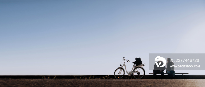 Morning bike ride along the city embankment. Silhouettes of a man and a woman on a bench against a blue sky. Copy space, space for text.