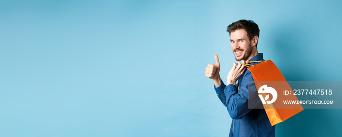 Cheerful shopper holding orange shopping bag on shoulder, turn around at camera with thumbs up, recommending store, blue background