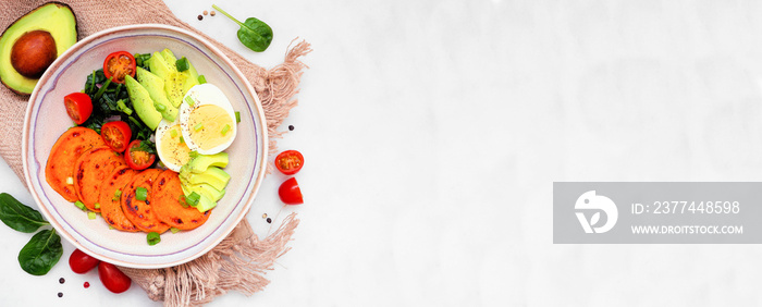 Nutritious breakfast bowl with sweet potato, egg, avocado and spinach. Overhead view table scene on a white marble banner background. Copy space.