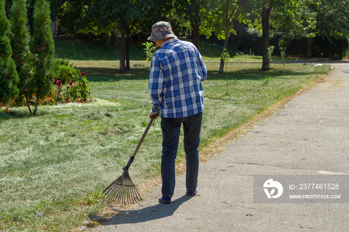 The janitor rakes dry leaves near the footpath. The janitor in a plaid shirt and jeans with a broom. The janitor in the hat cleans up garbage on the street.