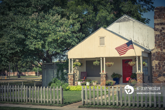 Vintage modest house with chimney in historic downtown district of Irving, Texas, USA. Classic wooden fence with well-groomed landscape, haning flower pots, big tree and proudly American flag waving