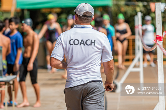 Back of male swimming coach walking on the poolside toward his young swimmers