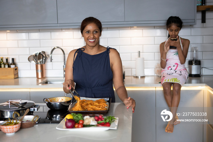 UK, London, Smiling�woman cooking in kitchen, daughter looking at smart phone