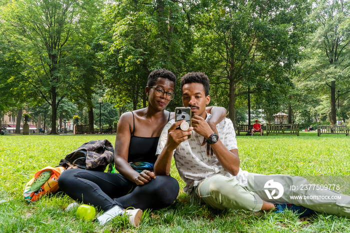 Couple sitting on lawn in park, looking at smart phone