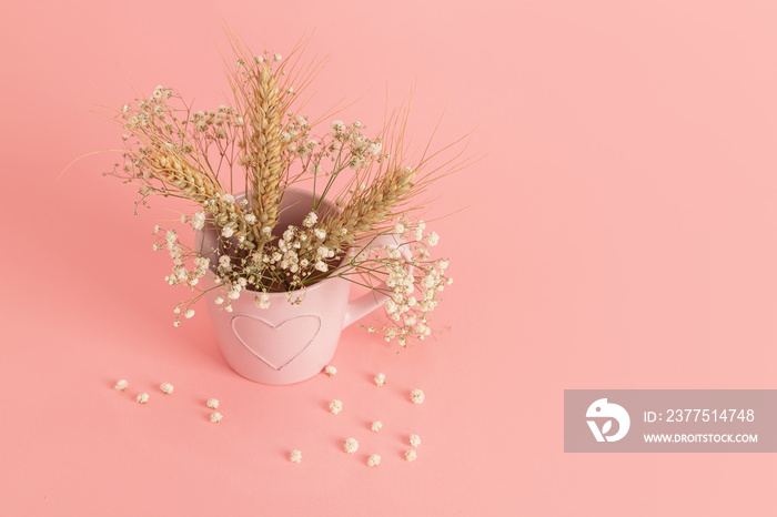 hite flowers of gypsophila and ears of wheat in a pink mug with a picture of a heart on a pink backg