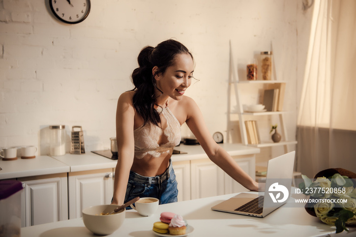 beautiful smiling young woman in lace bralette and jeans using laptop during breakfast at home