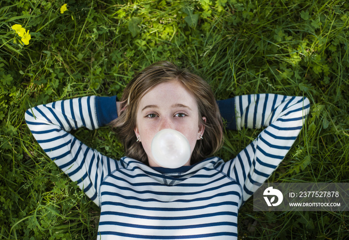 Portrait of girl with hands behind head blowing bubble gum while lying on field