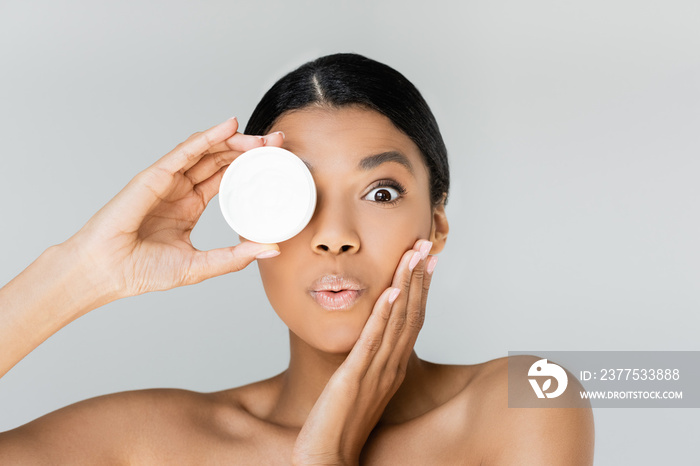 surprised african american woman covering eye while holding container with face cream isolated on gr