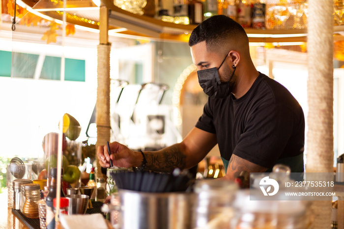 Young Latin American waiter working at the bar. He wears a face protection mask. Space for text.