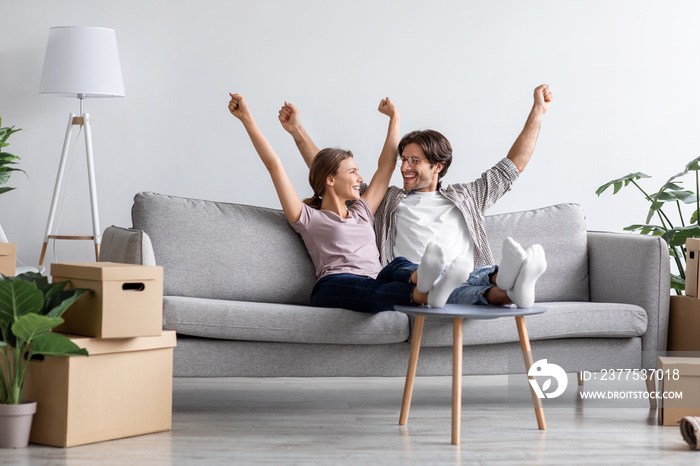 Happy excited european young husband and wife rejoice, raise their hands up on sofa in living room i
