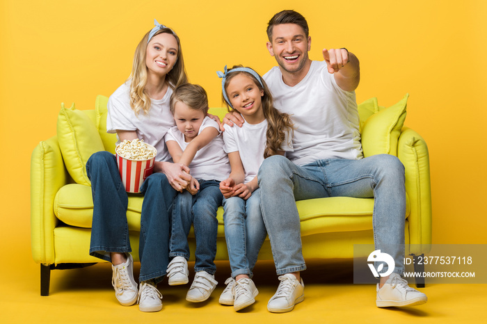 smiling parents and kids pointing and watching movie on sofa with popcorn bucket on yellow