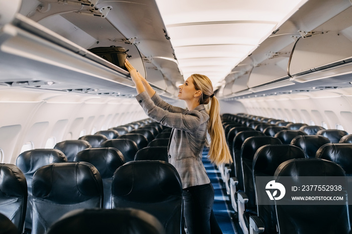 Young woman taking hand luggage out of airplane shelf.