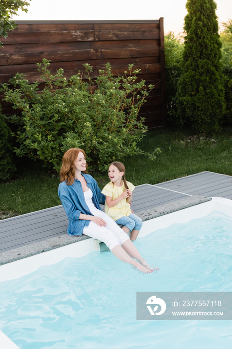 幸福的母女双腿坐在草坪附近的游泳池里的高角度视图