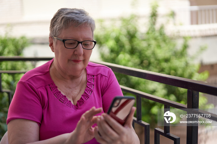 Older woman is sitting in a chair on her terrace at home. She is holding a red smartphone and typing