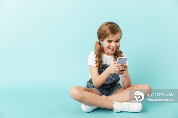 Happy young little girl posing isolated over blue wall background using mobile phone chatting.