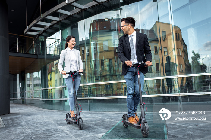 Two smiling business people driving electric scooter in front of modern business building going on w