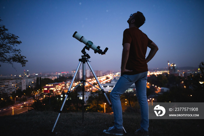 Astronomer with a telescope watching at the stars and Moon with blurred city lights in the backgroun