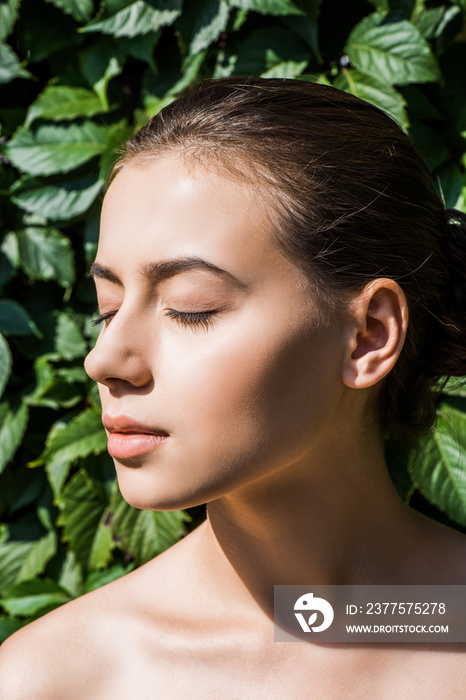 young beautiful woman with closed eyes against sunshine with green leaves at background