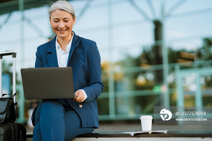 Grey asian woman working with laptop while sitting on bench by airport