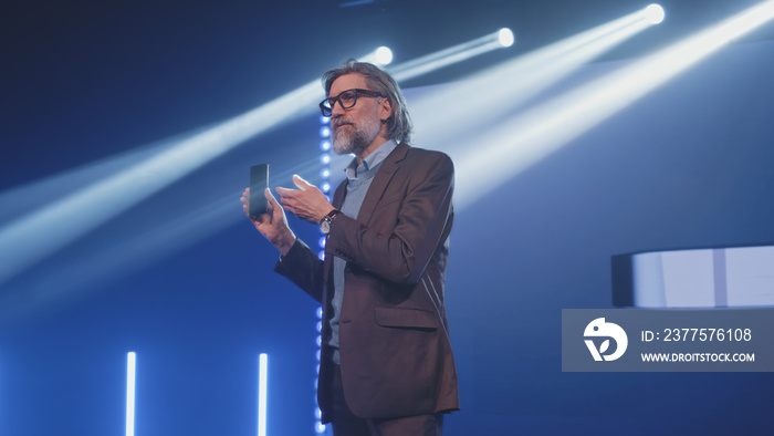 A male speaker on stage holding a new smartphone and talking about the characteristics, while standi