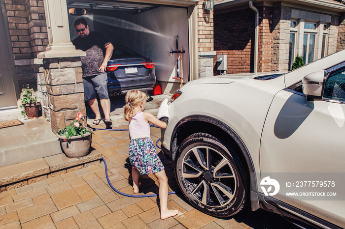 Cute preschool little Caucasian girl helping father wash car on driveway in front house on summer da