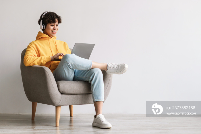 Happy young Asian man using laptop, wearing headphones while sitting in armchair against white wall,