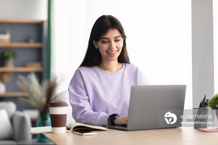 Cheerful millennial lady sitting at desk using laptop