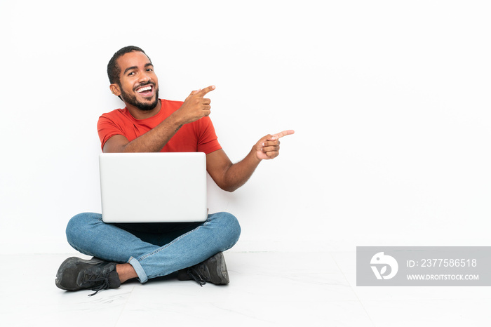 Young Ecuadorian man with a laptop sitting on the floor isolated on white background pointing finger