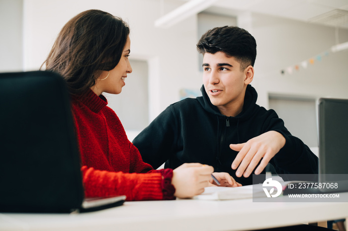 Smiling teenage friends discussing over book while sitting at desk in university