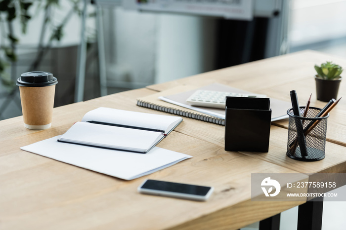 mobile phone with blank screen near empty notebook and paper cup on desk in office