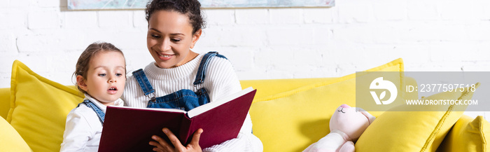 Panoramic shot of mother and daughter reading book on yellow couch at home