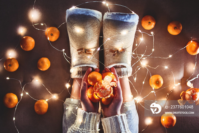 Ripe mandarins orange in hands, warm white christmas lights garland and female legs wearing warm fluffy soft winter slippers at cozy home at christmas eve. Top view