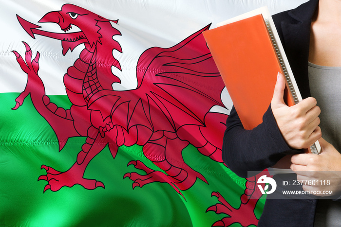 Learning Welsh language concept. Young woman standing with the Wales flag in the background. Teacher holding books, orange blank book cover.