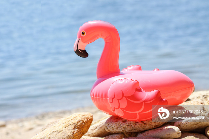 Inflatable little pink flamingo by the sea on  pebbles. Recreation and vacation concept, children’s game and fun. Summer relaxation on the beach. The sun is shining brightly, weather is great