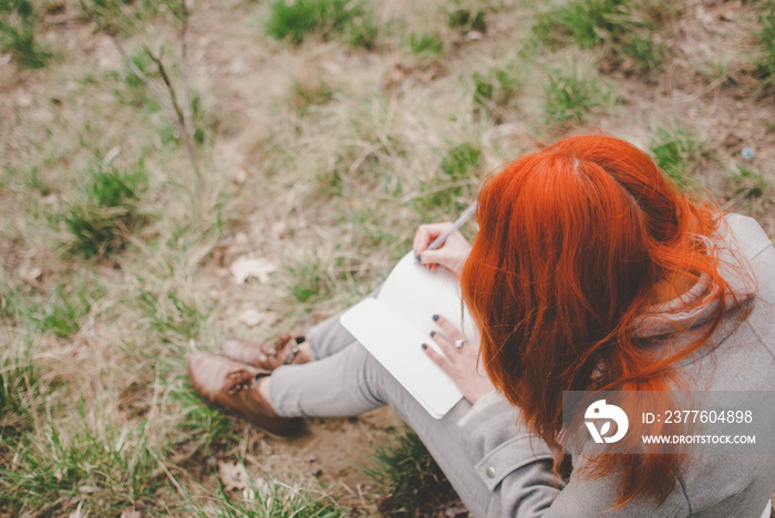 Redhead girl wearing a grey coat sitting on a green spring grass writing down her thoughts in the notebook. Pale hands and blank paper of the notebook. Concept of thinking, planning, and studying.