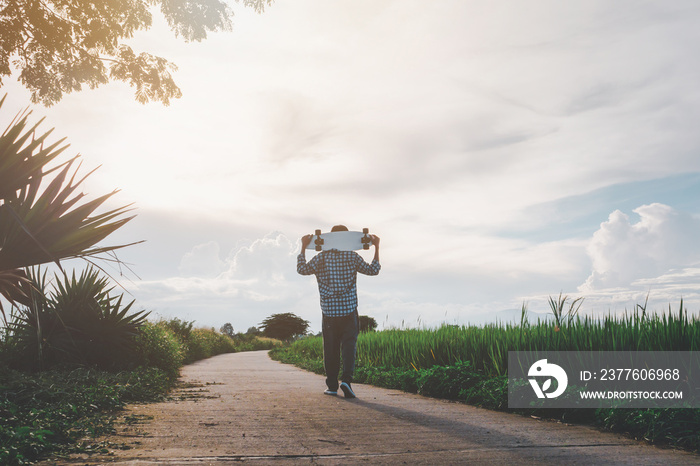 Young man stands with a skateboard behind his neck and looks over the evening fields.