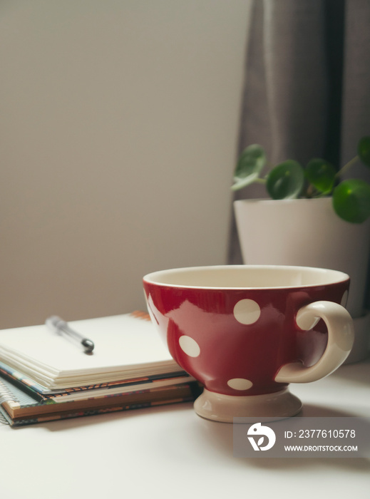 A mug of cappuccino sits on a white tabletop with an open notebook and a plant.