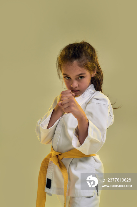 Portrait of little karate girl in white kimono with a yellow sash looking focused at camera, fighting, doing martial arts while standing isolated over yellow background