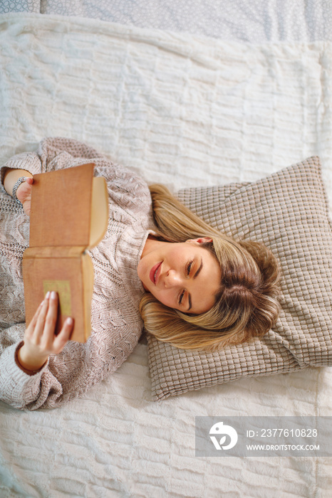 Young woman lies on her back in bed reading a book