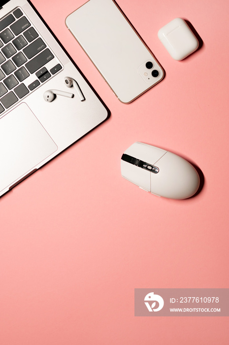 Overhead vertical flat lay photo with a silver grey laptop computer, modern smartphone, white plastic mouse, and wireless earphones with a charging case on a pink background.