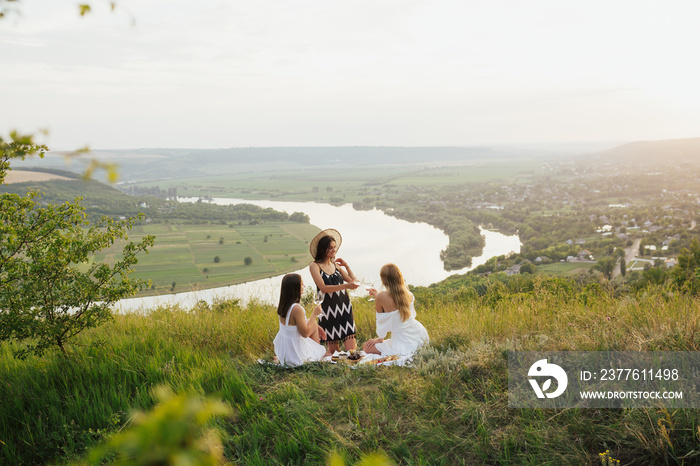 Three young beautiful women female friends are having picnic with wine, cheese and fresh fruits on the hill.